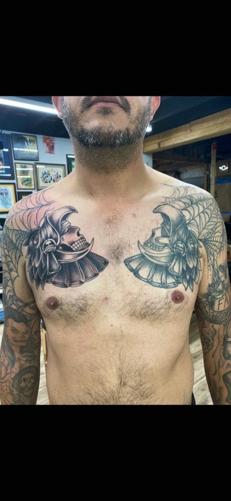Tattooed man with large black and gray skull and wing designs on his chest and colorful tattoos on arms.