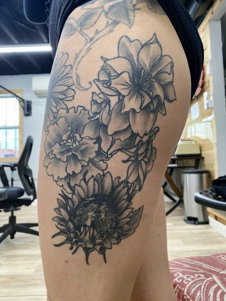 Detailed black and gray floral tattoo featuring a sunflower, lilies, and other flowers on thigh.