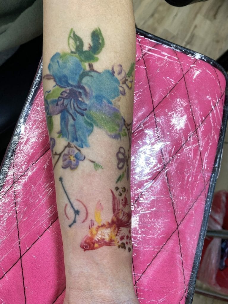 Colorful tattoo of blue flowers, a fish, and a small animal on a persons forearm.