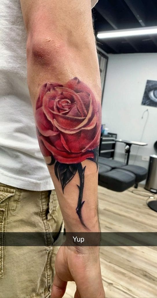 Detailed tattoo of a red rose with black thorns on a persons forearm.