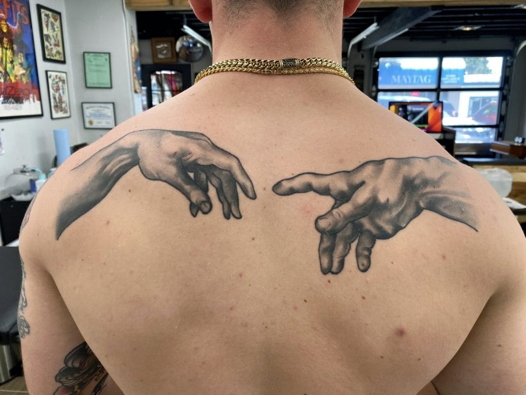 Tattoo of two realistic hands reaching towards each other on upper back and shoulders.