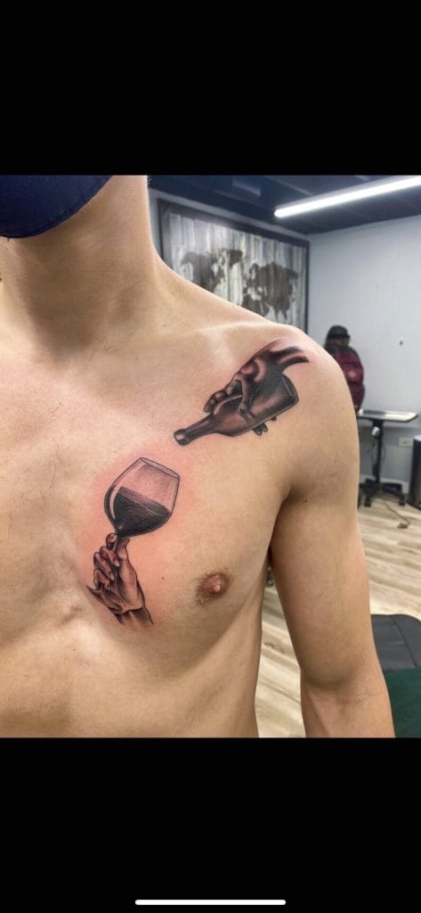 Tattoo of a hand holding a wine glass on the chest, with a bottle pouring wine onto the glass.