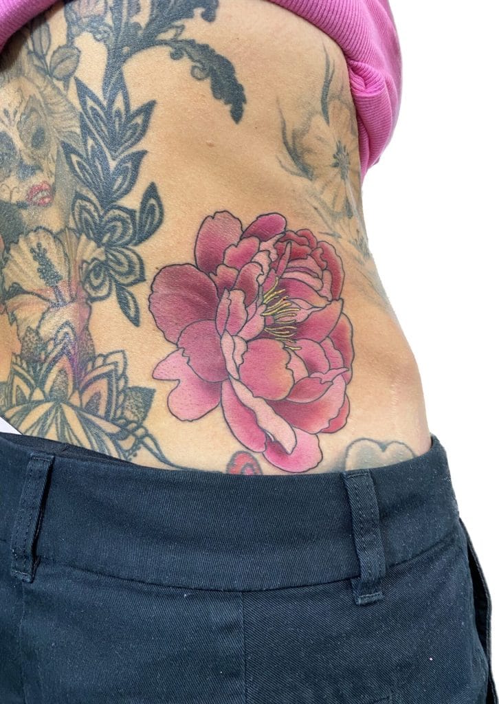 Pink and red peony tattoo on persons side waist, with other black and gray tattoos visible.
