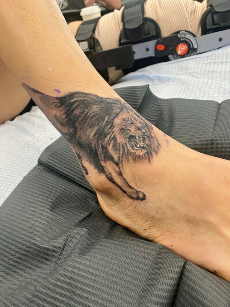 Tattoo of a fierce lion in mid-roar on a persons ankle and lower leg.