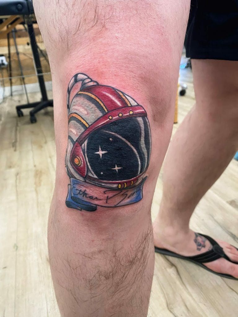 Colorful astronaut helmet tattoo with stars and a banner reading Earth I Shine on the knee.