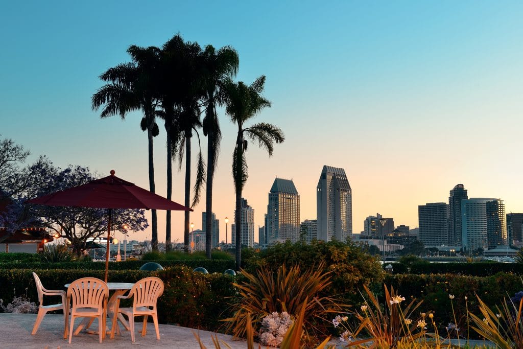 Plastic chairs and a table under a red umbrella overlook a garden with palm trees and the San Diego city skyline at sunset, just steps away from some of the city's iconic tattoo shops and rich San Diego tattoo history.