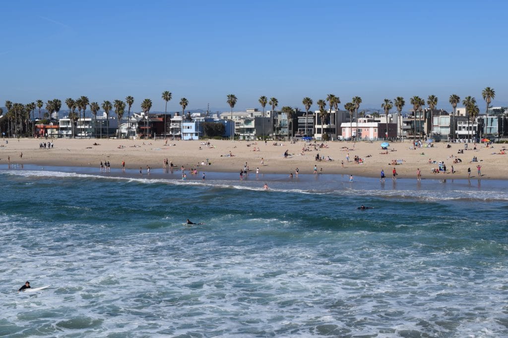 People relaxing and walking on a sandy San Diego beach with palm trees and buildings in the background; surfers ride the blue ocean under a clear sky at one of the city's top photo spots and tattoo-friendly beaches.