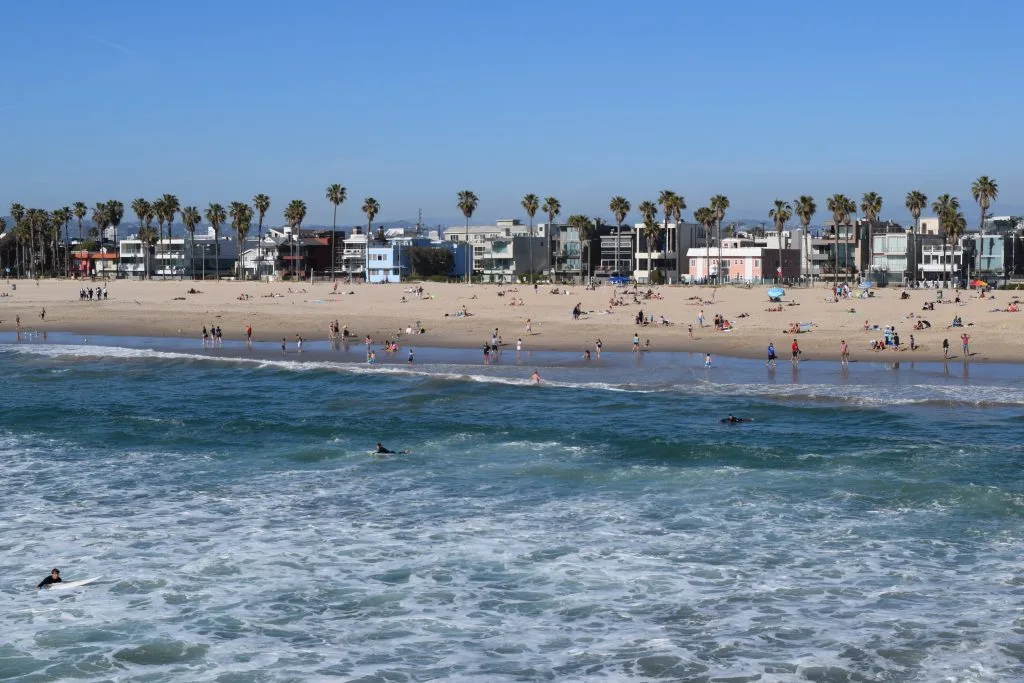 People relaxing and walking on a sandy San Diego beach with palm trees and buildings in the background; surfers ride the blue ocean under a clear sky at one of the city's top photo spots and tattoo-friendly beaches.