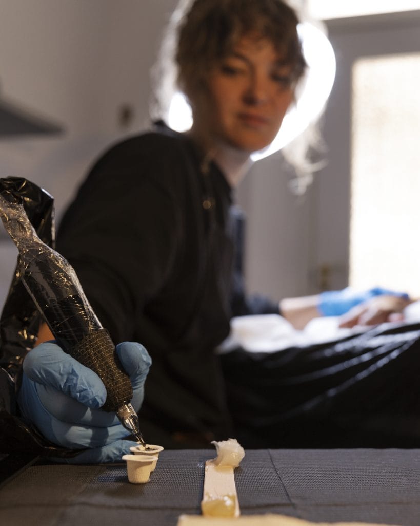A person wearing blue gloves dips a tattoo machine into ink, with tattoo supplies on a table in the foreground and the artist blurred in the background, highlighting the process of adding white ink highlights and their unique pros and cons.