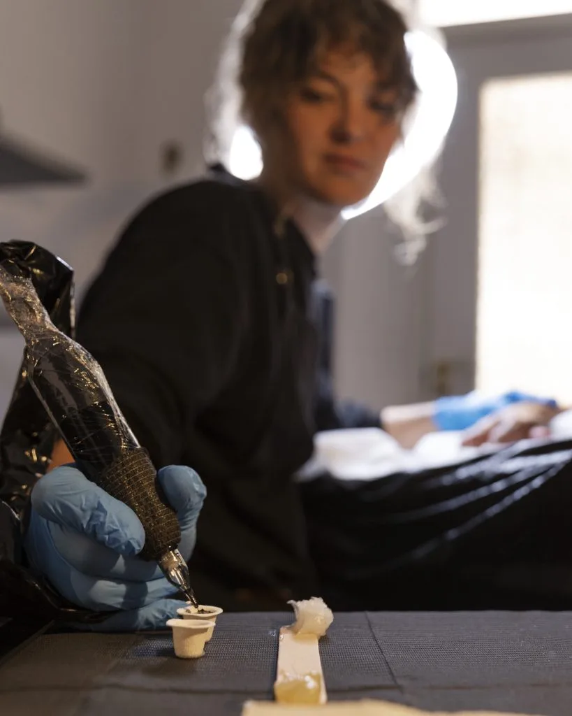 A person wearing blue gloves dips a tattoo machine into ink, with tattoo supplies on a table in the foreground and the artist blurred in the background, highlighting the process of adding white ink highlights and their unique pros and cons.