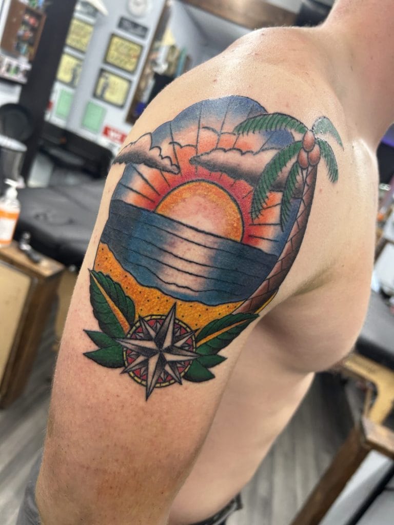 Colorful sunset over the ocean with a palm tree, leaves, and a compass tattoo on upper arm.