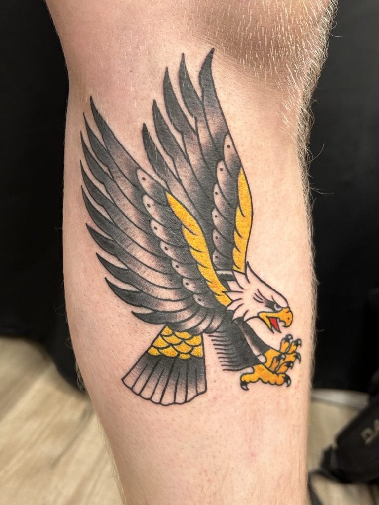 Detailed tattoo of a fierce eagle with outstretched wings, claws, and beak, in black, gray, yellow, and orange.