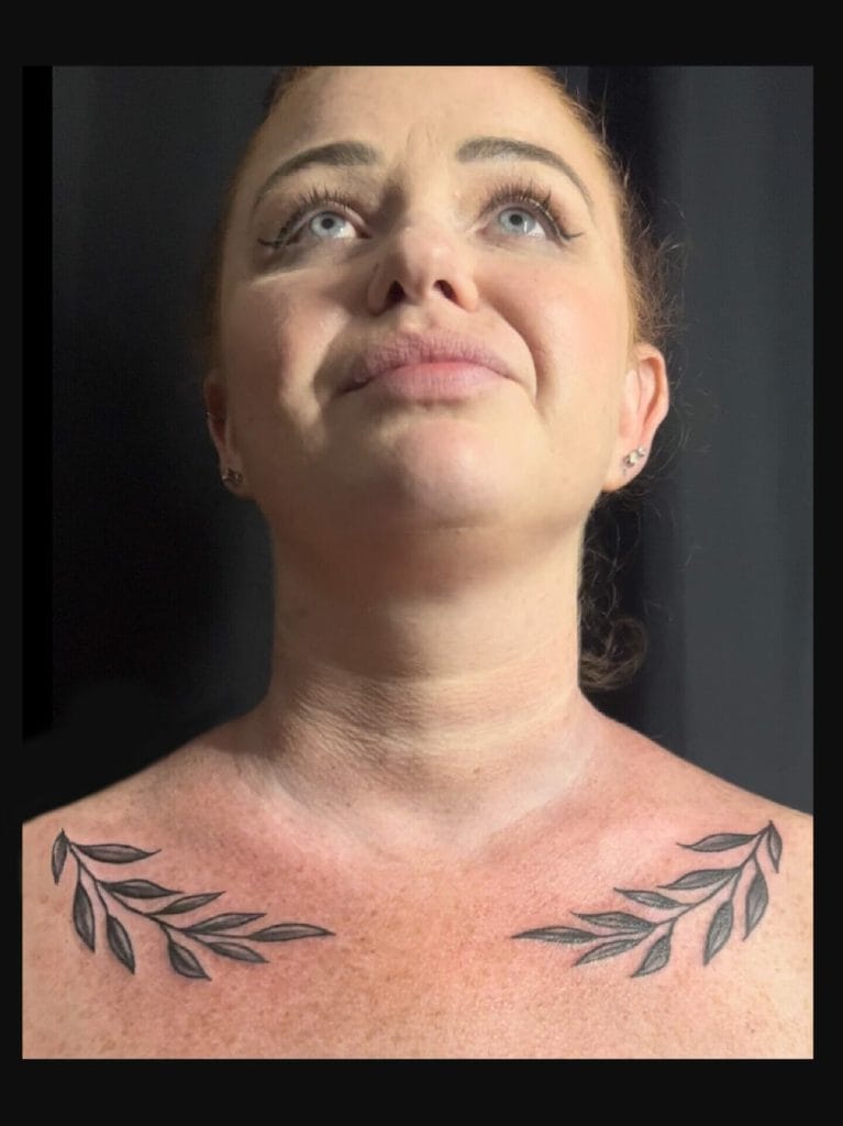 Woman with light skin and blue eyes, looking upward, with tattoos of leaves on her shoulders.