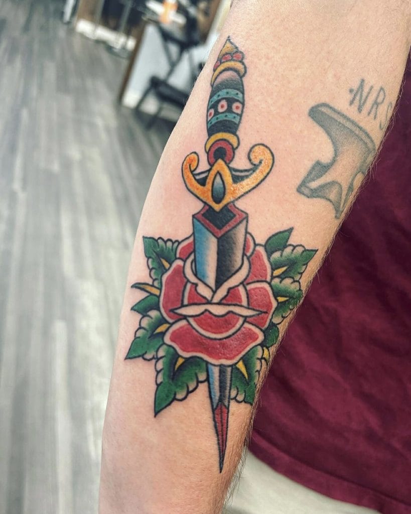 Colorful dagger piercing a red rose with green leaves tattoo on forearm.