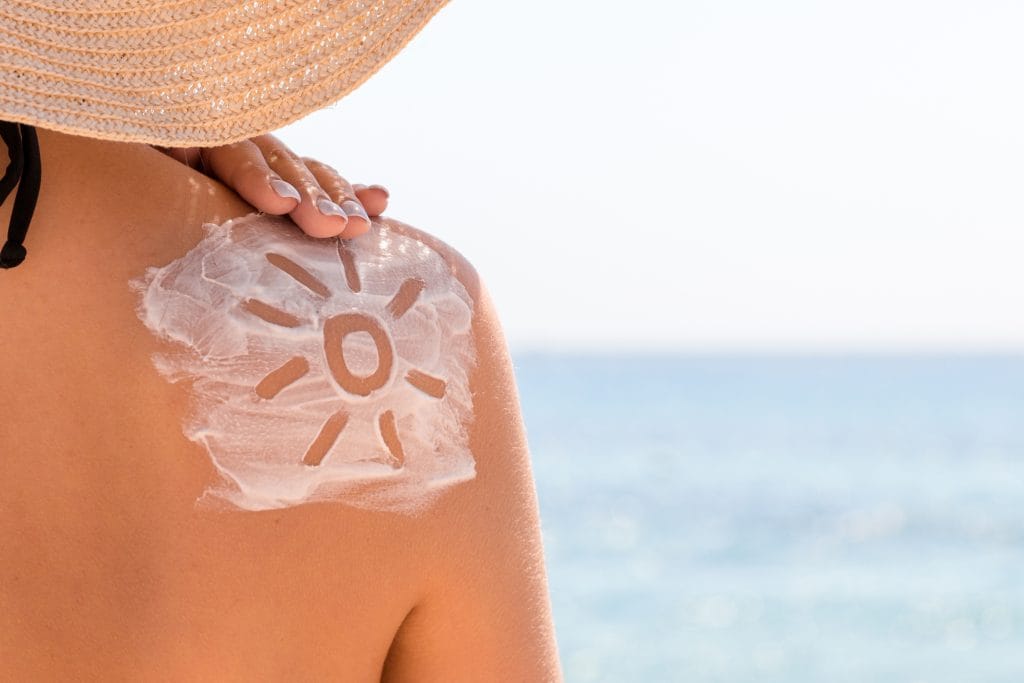 A person wearing a sunhat at the beach in San Diego displays a sun-shaped sunscreen design on their shoulder, with the sea and coastal climate visible in the background.