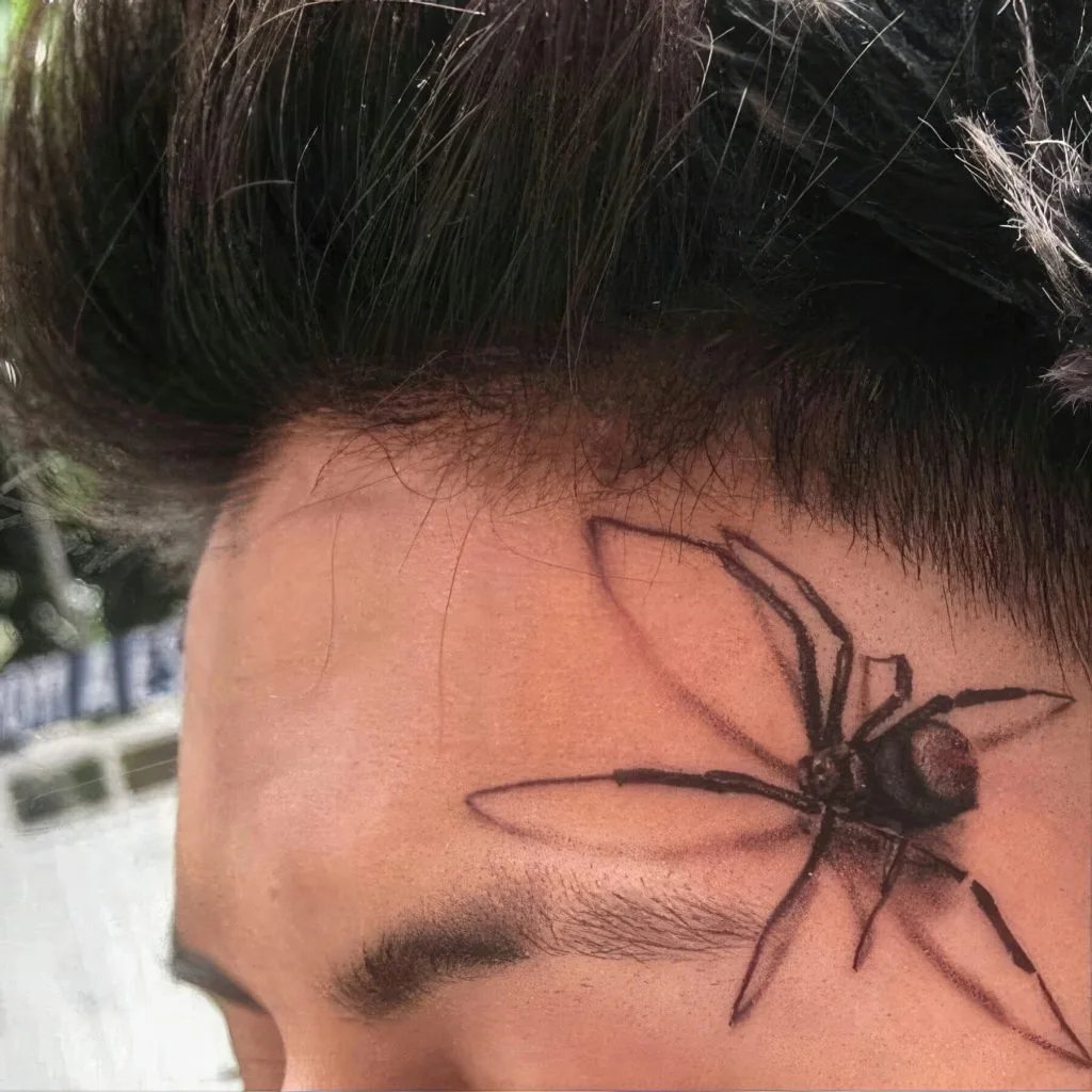 Tattoo of a black spider on the persons forehead near the hairline.