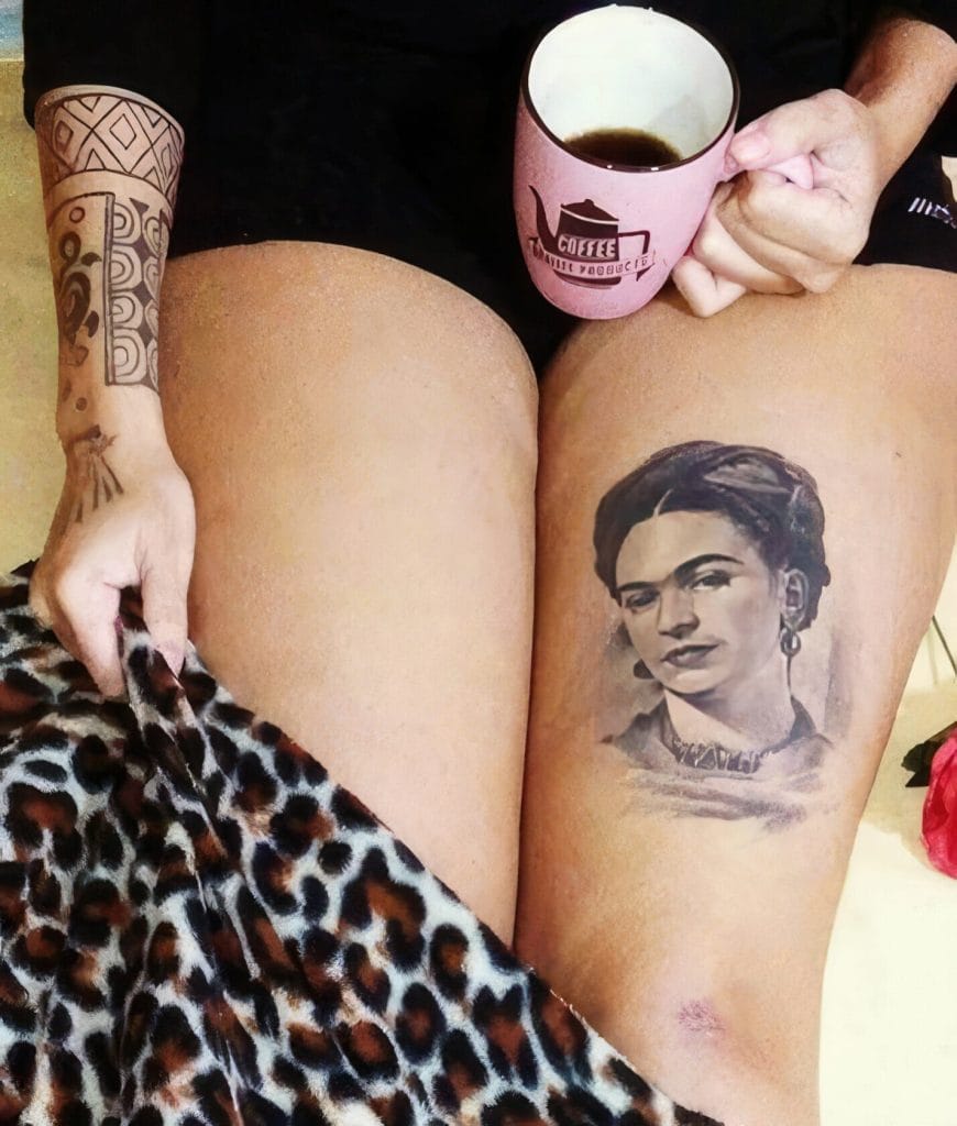Person with a tattoo of Frida Kahlo on thigh, holding a coffee mug, wearing leopard print fabric.