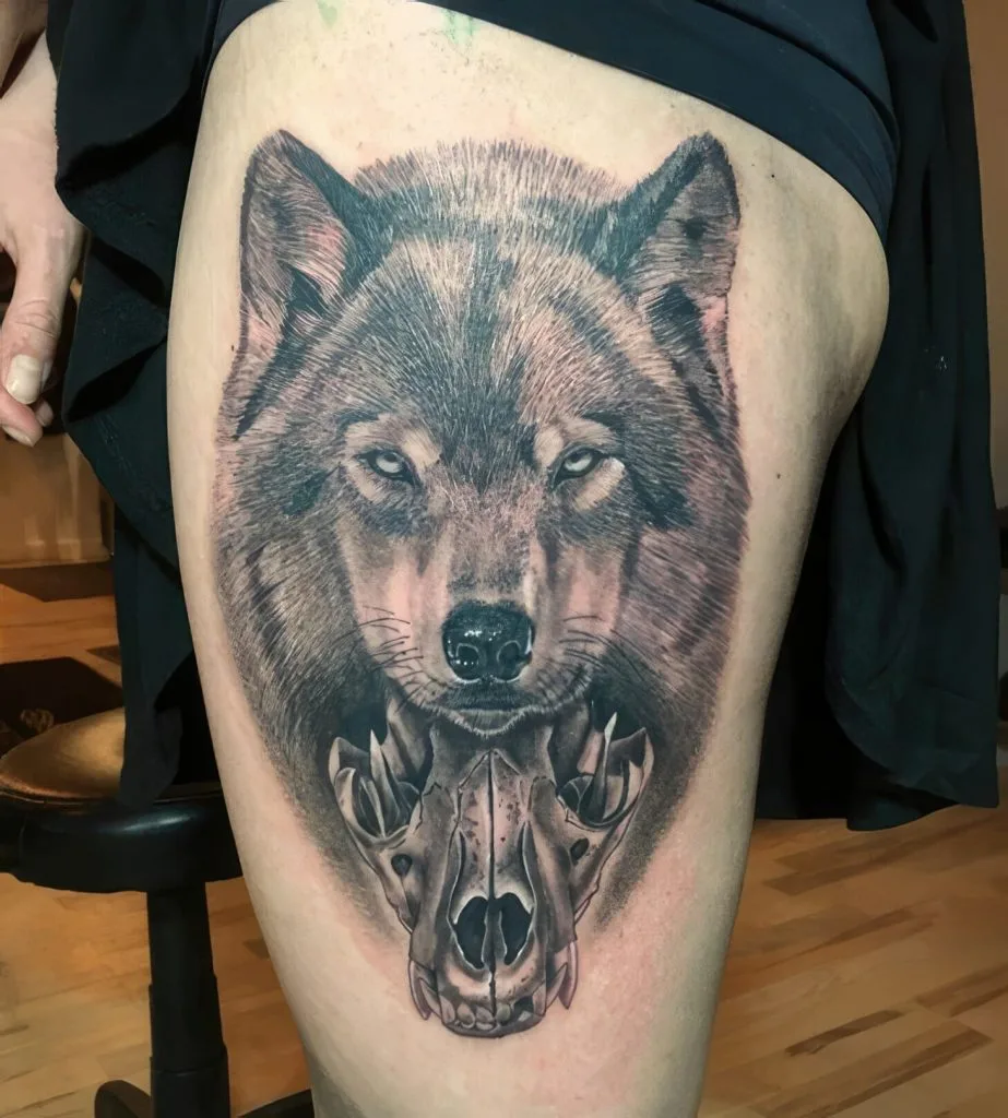 Detailed black and gray wolf head tattoo with a skull beneath, on persons thigh.