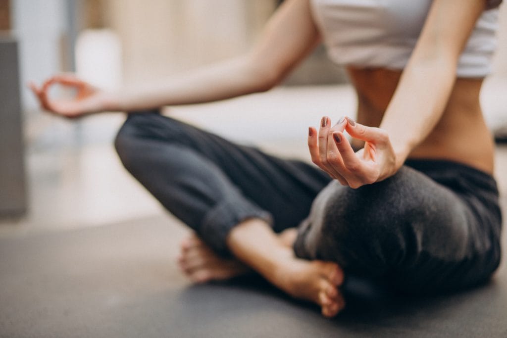 Person sitting cross-legged on a mat, wearing a white top and dark pants, with hands resting on knees in a meditation pose—exploring natural alternatives to numbing creams for relaxation and calm.