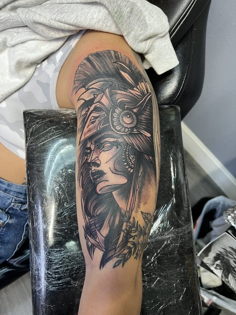 Black and gray tattoo of a woman wearing a feathered headdress with leaves below.