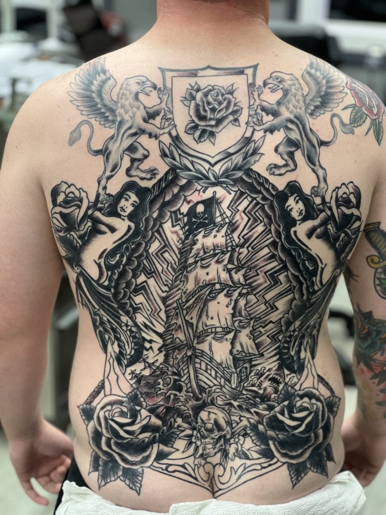 Detailed black and gray tattoo of a sailing ship, roses, skull, and mythical creatures on persons back.