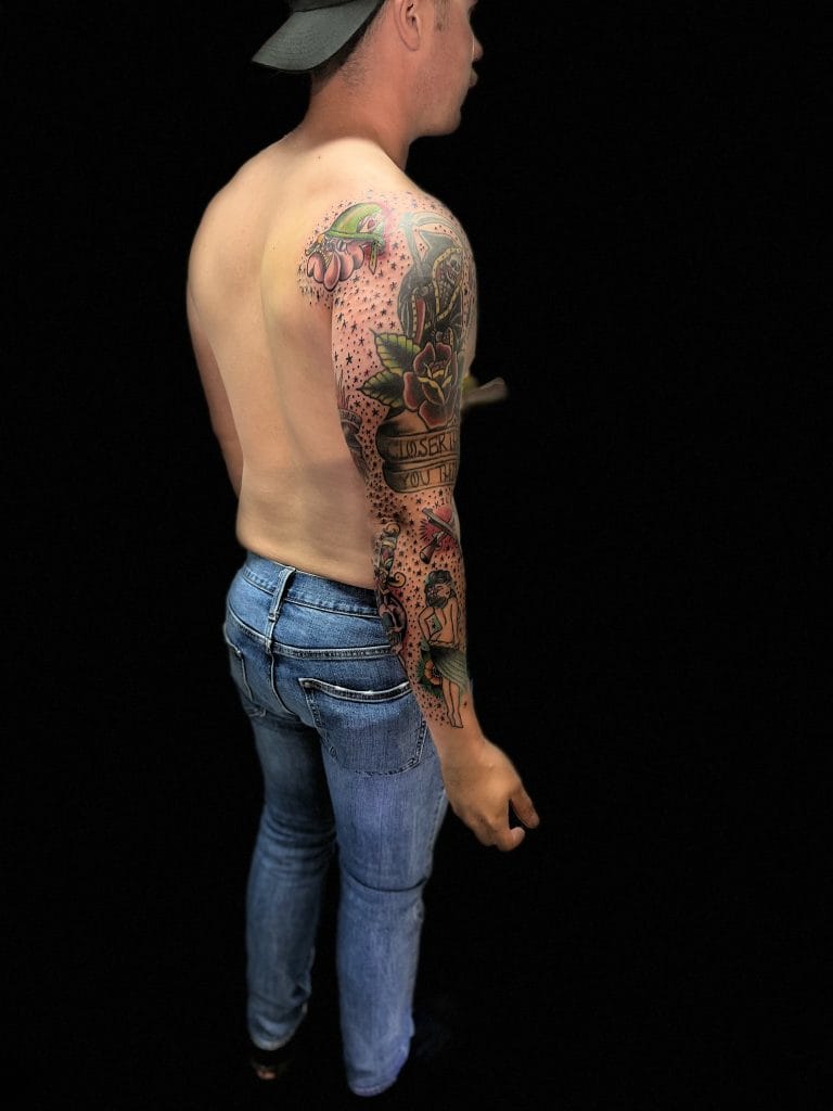 Side view of a shirtless man with colorful tattoos on his left arm, wearing a gray cap and blue jeans.