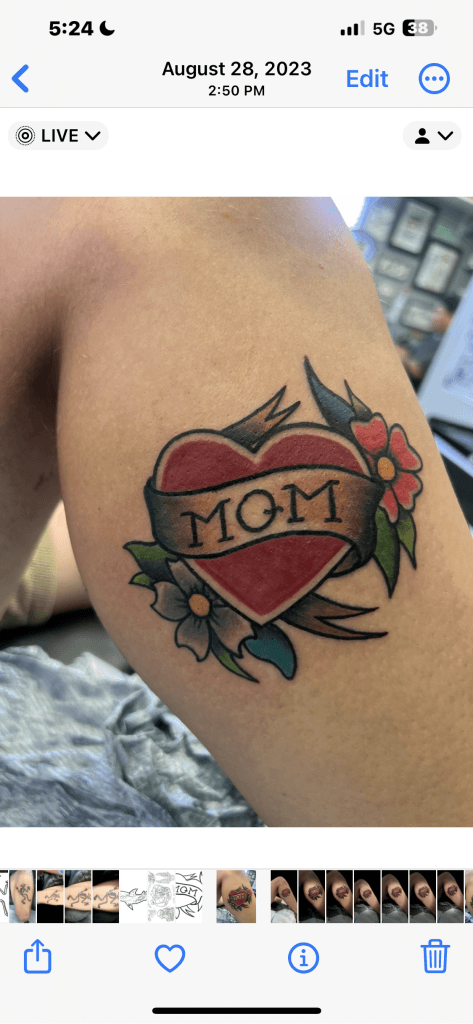Red heart tattoo with MOM banner, surrounded by colorful flowers and leaves.