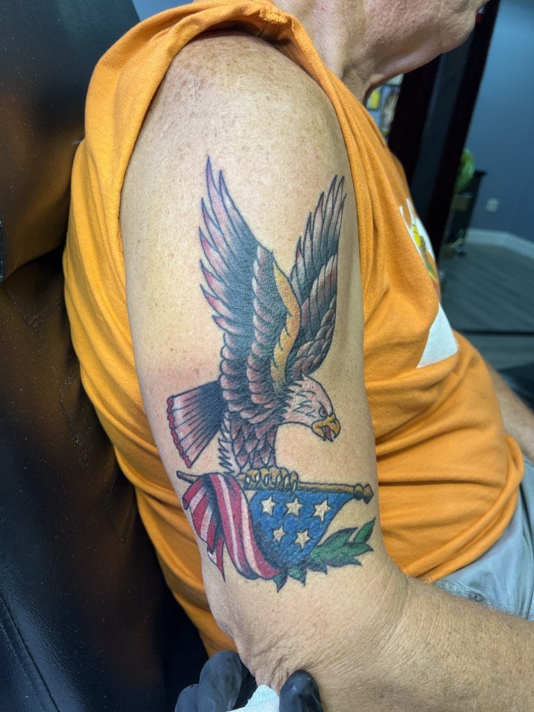 Tattoo of an eagle holding an American flag with stars and stripes on the upper arm.
