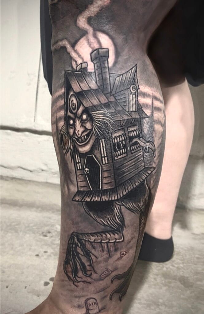 A detailed black and gray tattoo of a haunted house with a sinister face, on a persons thigh.